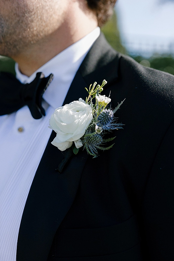 Groom boutonniere with a white rose boutonniere and blue thistle pinned to a black tuxedo lapel against soft outdoor greenery and sky