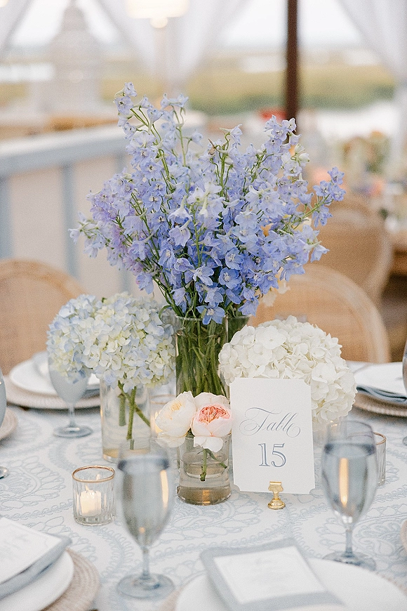 Reception tablescape with wedding table centerpiece of blue delphinium and white hydrangeas in glass vases, candlelit under a tent canopy