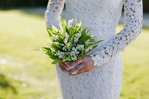 Bridal bouquet of lily of the valley bouquet held by a bride in long lace sleeves, wedding ring visible in sunlit lawn greenery