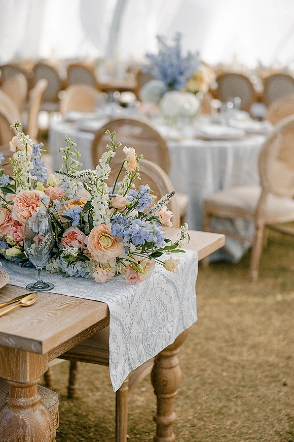 Reception tablescape with a pastel wedding centerpiece on a patterned runner, gold flatware and champagne flute on wooden table under tent on lawn