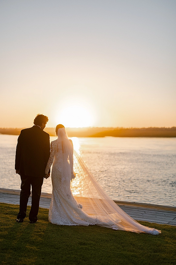 Couple portrait of bride and groom silhouette holding hands on a lakeside dock at sunset, her long veil trailing behind