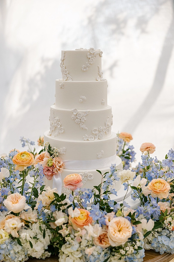 Wedding cake with white fondant tiers, sugar flowers and satin ribbon on a stand, framed by pastel florals against draped backdrop