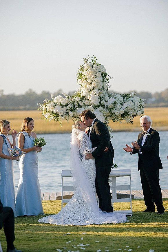 Ceremony kiss as bride in lace dress and veil kisses groom in tuxedo before a white floral cross altar by the waterfront lawn