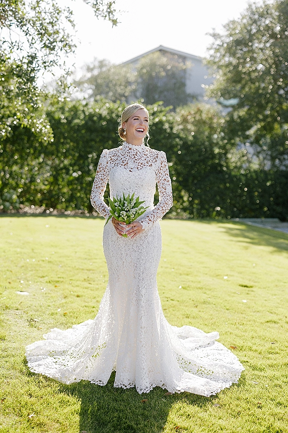 Bridal portrait of a bride in a lace long sleeve wedding dress holding a white and green bouquet on a sunlit garden lawn with hedges