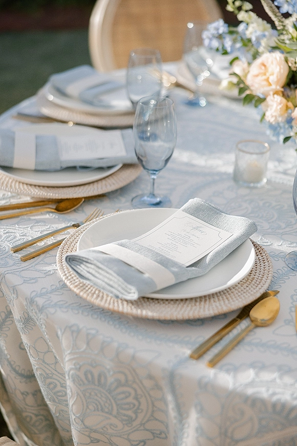Reception tablescape with outdoor wedding table setting on a lawn, featuring patterned cloth, rattan chargers, gold flatware, blue stemware, and florals