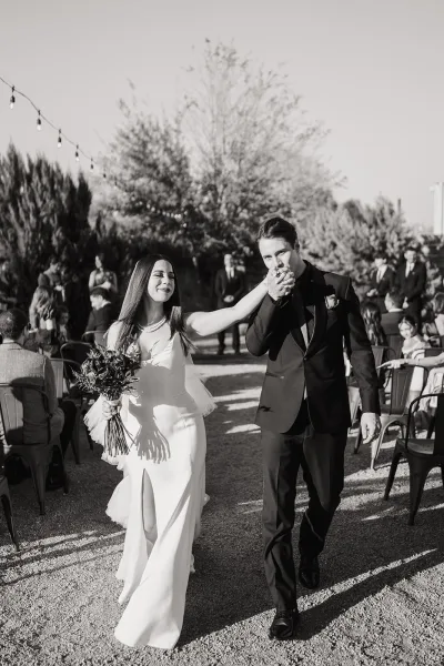 Wedding recessional as bride and groom walk the aisle hand in hand, groom kissing her hand beneath string lights in a garden setting