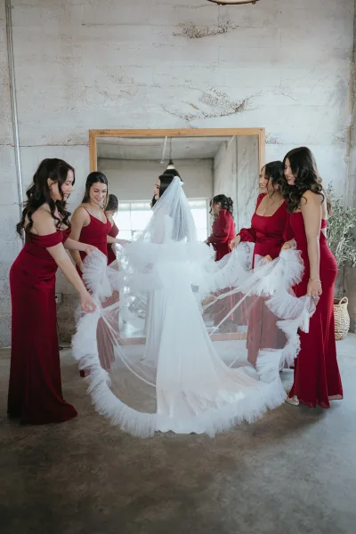 Bride with bridesmaids holding her cathedral veil as it fans out behind her in a minimal loft, window light reflecting in a floor mirror