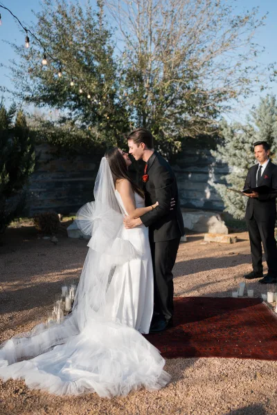 Ceremony kiss at an outdoor wedding ceremony as bride in long veil and groom in black suit share a kiss on a candle-lined rug aisle in a garden