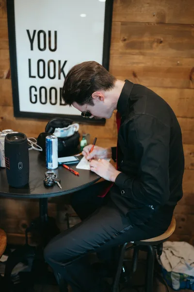 Groom writing vows in a wedding vows notebook, seated at a table in sunglasses and red tie against a rustic wood plank wall
