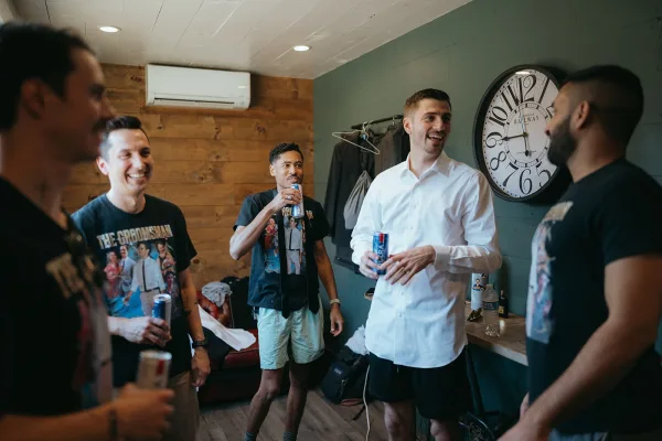 Groom getting ready with groomsmen, buttoning a white dress shirt and tie as beer cans and suit jackets sit in a small room