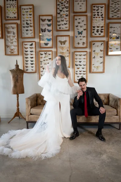 Couple portrait with bride in strapless gown and long veil holding groom’s hand by a tufted sofa in a butterfly art gallery space
