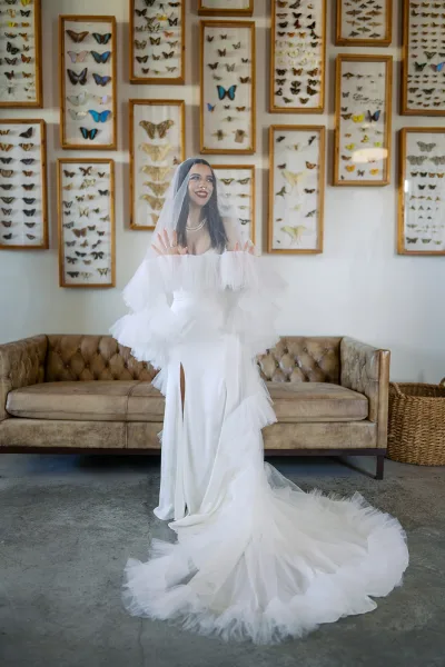 Bridal portrait of a bride in veil, smiling in a tulle sleeve wedding dress with pearl necklace and long train beside butterfly wall art and tufted sofa