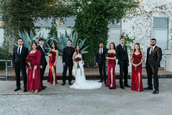 Wedding party portrait with bridesmaids in red dresses and groomsmen in black suits beside an ivy-covered industrial wall, bride’s train flowing