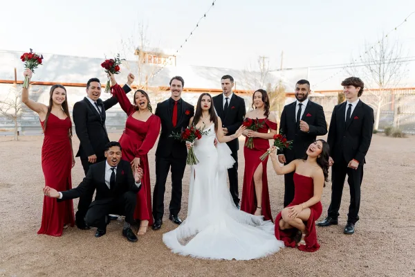 Wedding party portrait with bridesmaids in red and groomsmen in black suits cheering around the bride and groom under string lights outdoors
