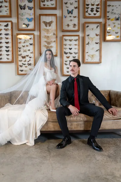 Couple portrait of bride and groom seated on a tufted sofa, her cathedral veil flowing, his red tie, with butterfly wall art behind