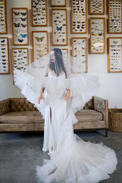 Bridal portrait of a bride in a spaghetti strap gown with tulle veil over her face, standing by framed butterfly displays and a tufted sofa indoors