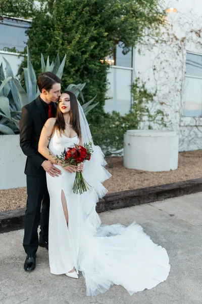 Couple portrait of groom kissing bride’s forehead as she holds a red bouquet, cathedral veil flowing beside ivy wall and modern planters