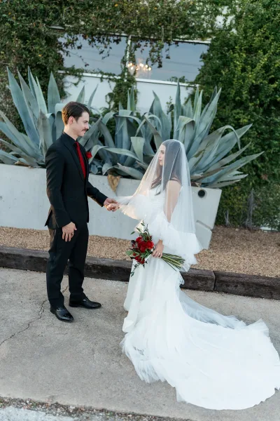 Couple portrait of bride and groom holding hands, veil over her face with red rose bouquet, beside agave plants and concrete wall outdoors