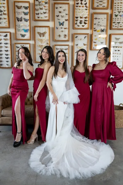 Bride with bridesmaids in burgundy dresses posing indoors, bride in white veil and pearl necklace before butterfly wall art backdrop