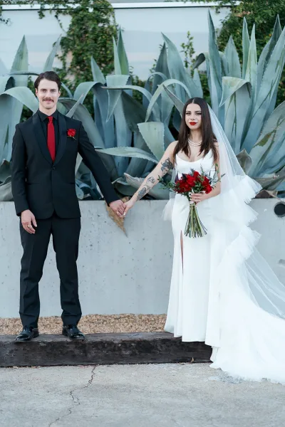 Couple portrait of bride and groom holding hands, bride in strapless dress with veil and red rose bouquet by agave greenery wall