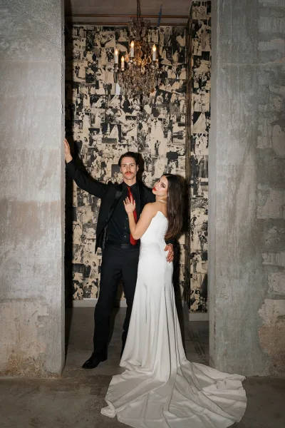 Couple portrait of bride in a strapless wedding dress with train and pearl necklace beside groom in black suit and red tie in a concrete hallway doorway under a chandelier