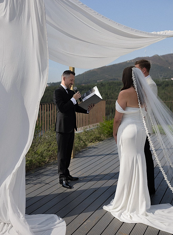 Wedding vows exchanged as an officiant reads on a microphone beneath a white draped arch, bride’s lace veil on a mountain deck