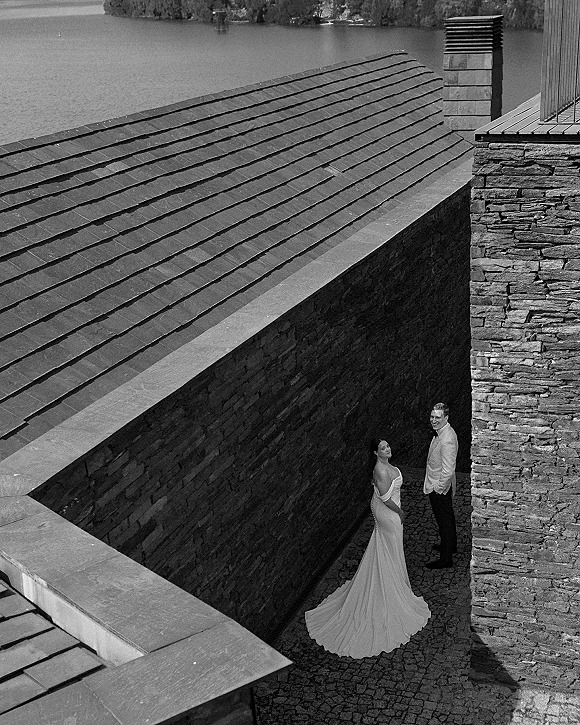 Couple portrait in a black and white wedding portrait, bride in strapless gown with long train beside groom in white jacket by stone wall and cobblestones