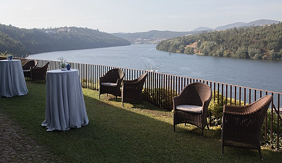 Cocktail hour setup with blue tablecloth high-top tables, small floral vases, and wicker lounge chairs overlooking a river and mountains
