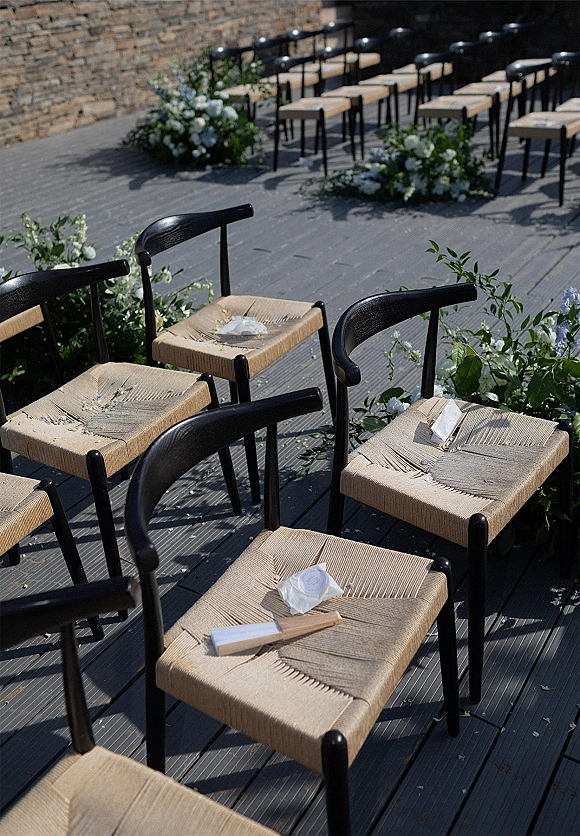 Ceremony seating with outdoor ceremony chairs lined on a wood deck, black woven seats, programs, rose petals, and white-blue florals by a stone wall