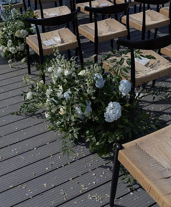 Ceremony aisle decor with aisle floral ground arrangements of blue and white blooms and greenery garlands beside black woven chairs on a wood deck floor