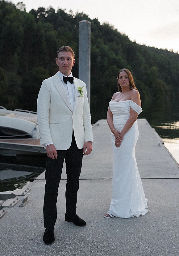 Couple portrait of bride in an off-the-shoulder wedding dress and groom in a white tuxedo jacket on a dock by the lake and boat