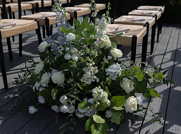 Ceremony aisle flowers with wedding aisle florals in white roses, light blue blooms and greenery lining a wood deck aisle with woven chairs