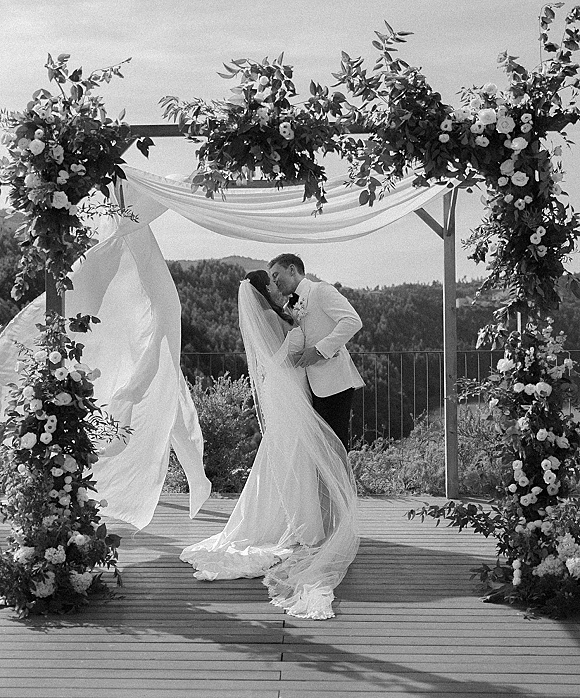 Wedding kiss under a floral arch with draped fabric, bride in long veil and gown, groom in tuxedo on deck with forested hills