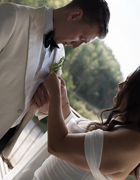 Couple portrait of bride and groom close up as she adjusts his boutonniere, his white tuxedo jacket and bow tie on a green balcony in daylight