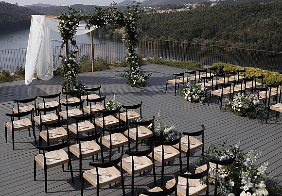 Outdoor ceremony setup with lakeview wedding ceremony seating, a wood arch draped in white fabric and greenery on a deck by the lake