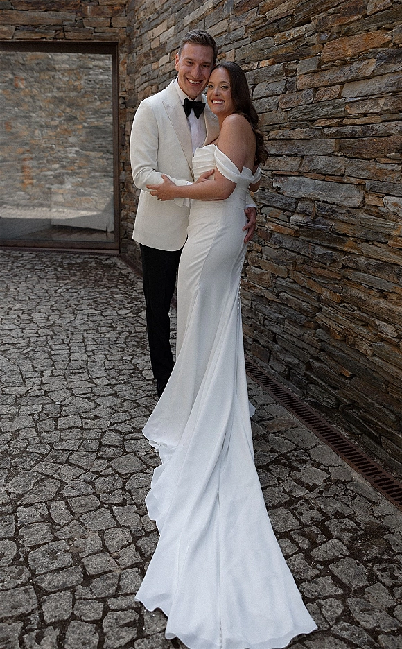 Couple portrait of bride and groom hugging, her strapless gown with long train beside a stone wall on a cobblestone walkway