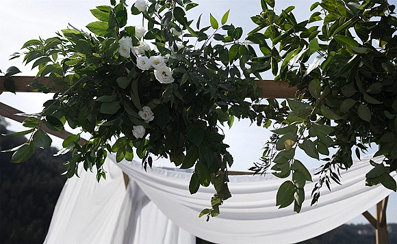 Wedding ceremony arch with greenery wedding arch garland, white flowers and flowing fabric drape on a wooden frame against hills and sky