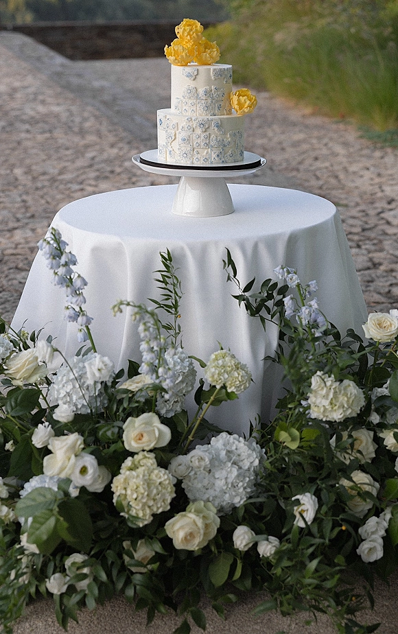 Wedding cake with a two tier wedding cake in blue and white tile fondant, topped with yellow sugar flowers on a white table outdoors