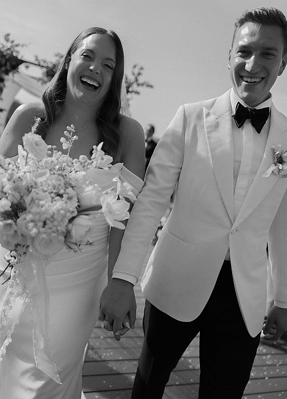 Couple portrait in a black and white wedding photo, bride and groom laughing hand-in-hand on a wooden deck with guests behind