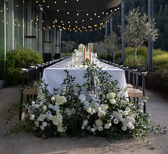 Wedding tablescape with long banquet table decor, white roses and hydrangea runner, taper candles, glassware, and bistro lights on a covered patio
