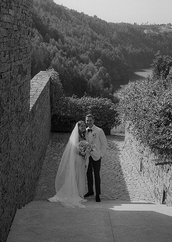 Couple portrait in black and white wedding portrait style, bride in gown and veil holding bouquet beside groom in tux on stone steps by lake