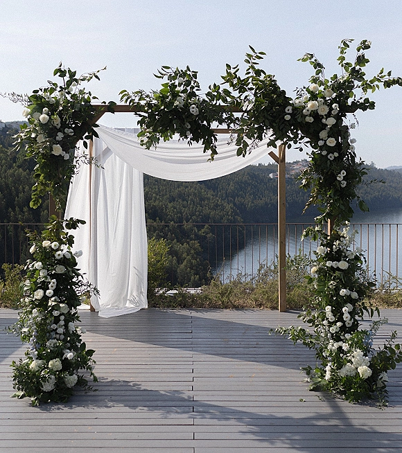 Wedding ceremony arch with white draping fabric and white rose floral clusters on a deck overlooking a lake and forested hills