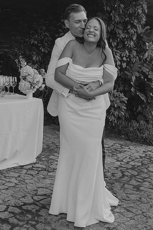 Couple portrait of bride and groom hug as he kisses her cheek, off-shoulder dress and bow tie on a cobblestone garden patio