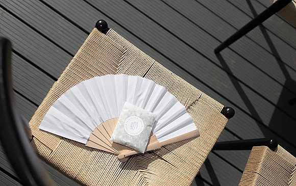 Wedding favor ceremony hand fan in a favor packet resting on a woven chair, with sunlit shadows across the wood deck floor