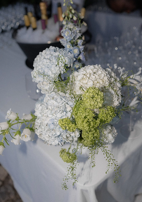 Wedding centerpiece with hydrangea centerpiece blooms and greenery in bud vases beside wine bottles on a white tablecloth with blurred glassware