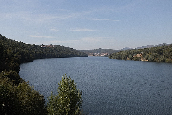 Lake landscape with a wide lake view of calm water, forested shoreline, distant town, mountains, and blue sky beyond