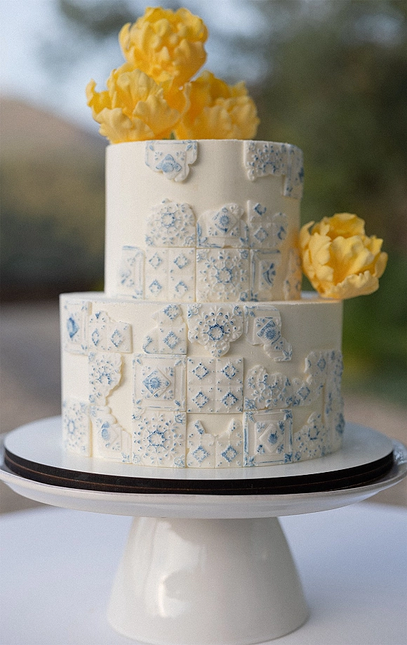 Wedding cake with blue and white tile-pattern fondant on a cake stand, topped with yellow flowers against blurred outdoor greenery