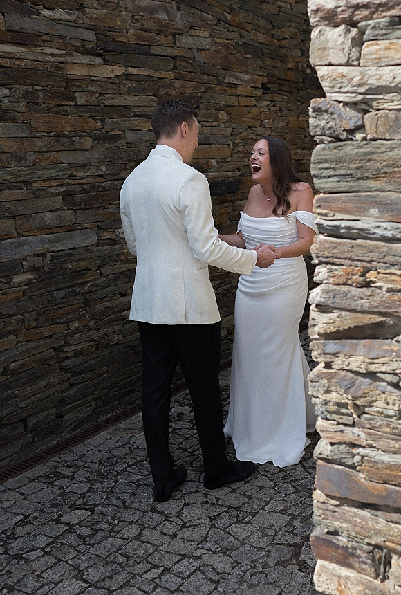 First look moment as bride and groom holding hands, bride in strapless gown laughing beside a stone wall on cobblestones outdoors