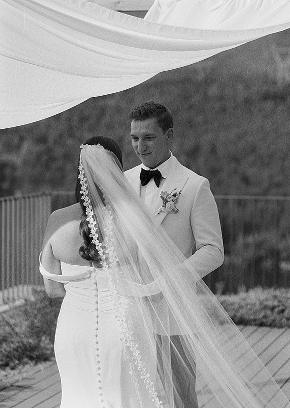 Wedding ceremony moment as bride and groom exchange vows holding hands under a draped canopy on a wooden deck, lace veil trailing behind