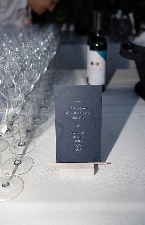 Wedding bar menu card on a stand with white ink typography beside wine bottle and champagne flutes on a linen bar table with bartender behind